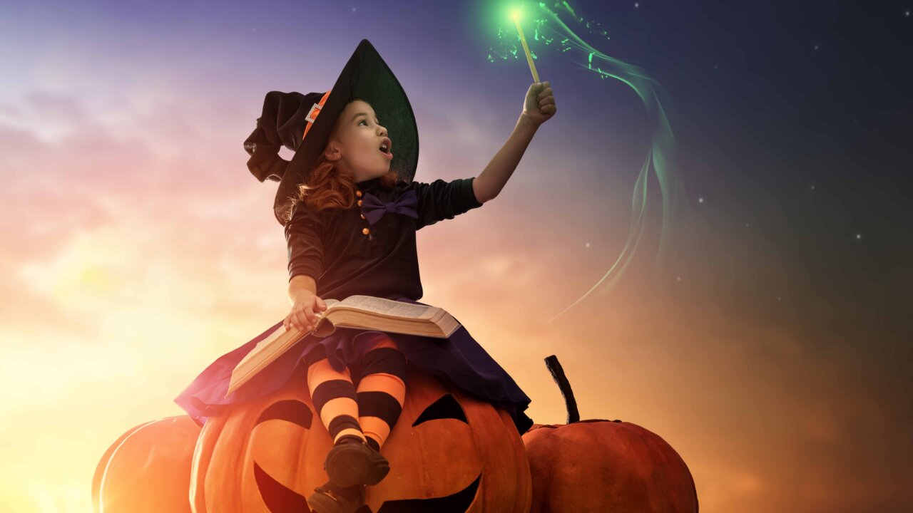 Cute cheerful little witch with a magic wand and book of spells. Beautiful child girl in witch costume sitting on the big pumpkin, conjuring and laughing.