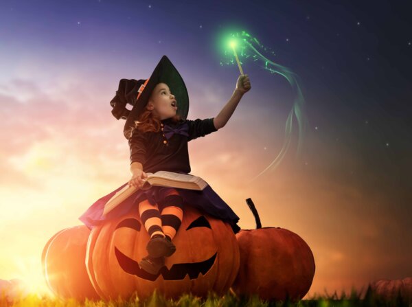 Cute cheerful little witch with a magic wand and book of spells. Beautiful child girl in witch costume sitting on the big pumpkin, conjuring and laughing.